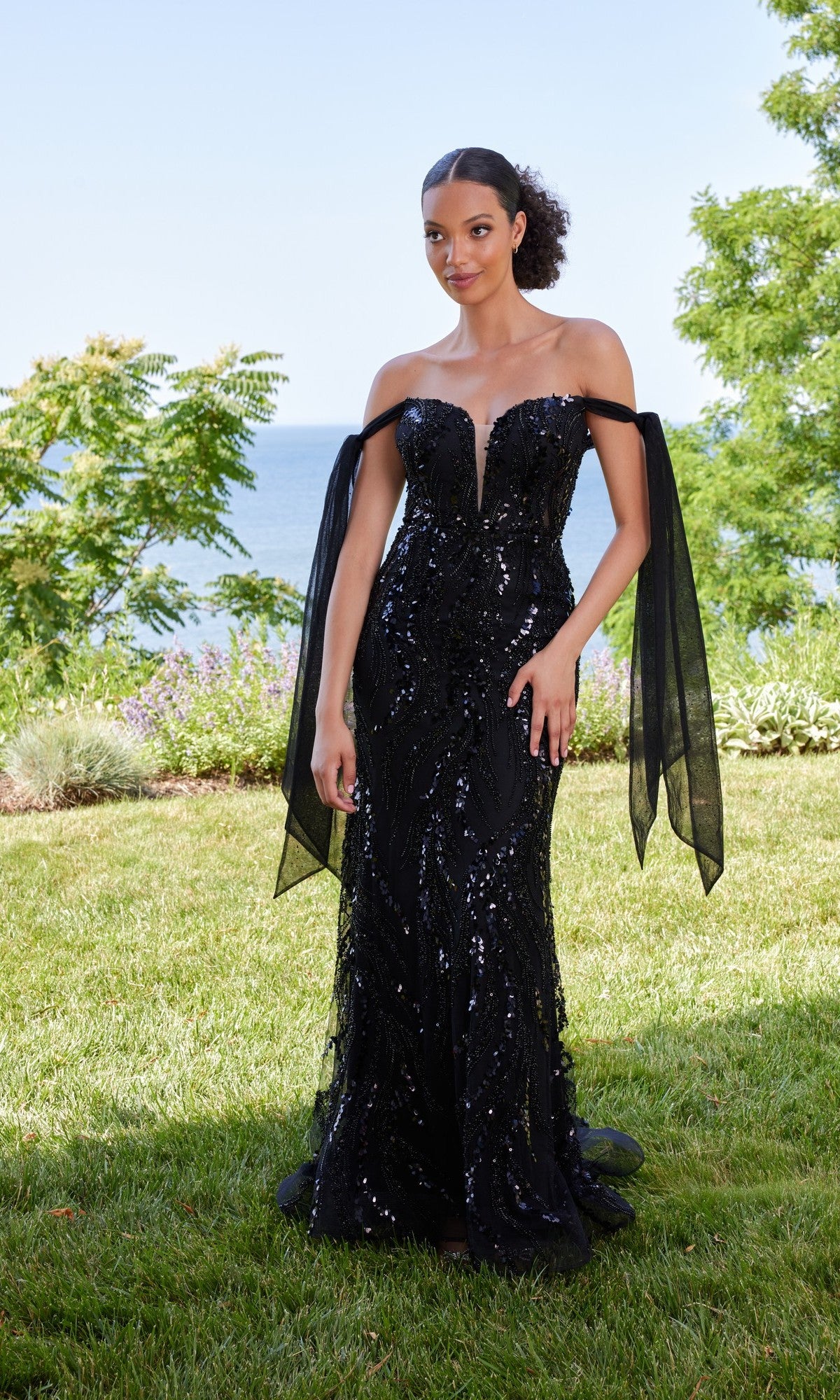 Outdoors on grass, a woman wears the Morilee 49166 long black prom dress with off-the-shoulder sheer ribbon sleeves and sparkling embellishments; trees and water appear in the background.